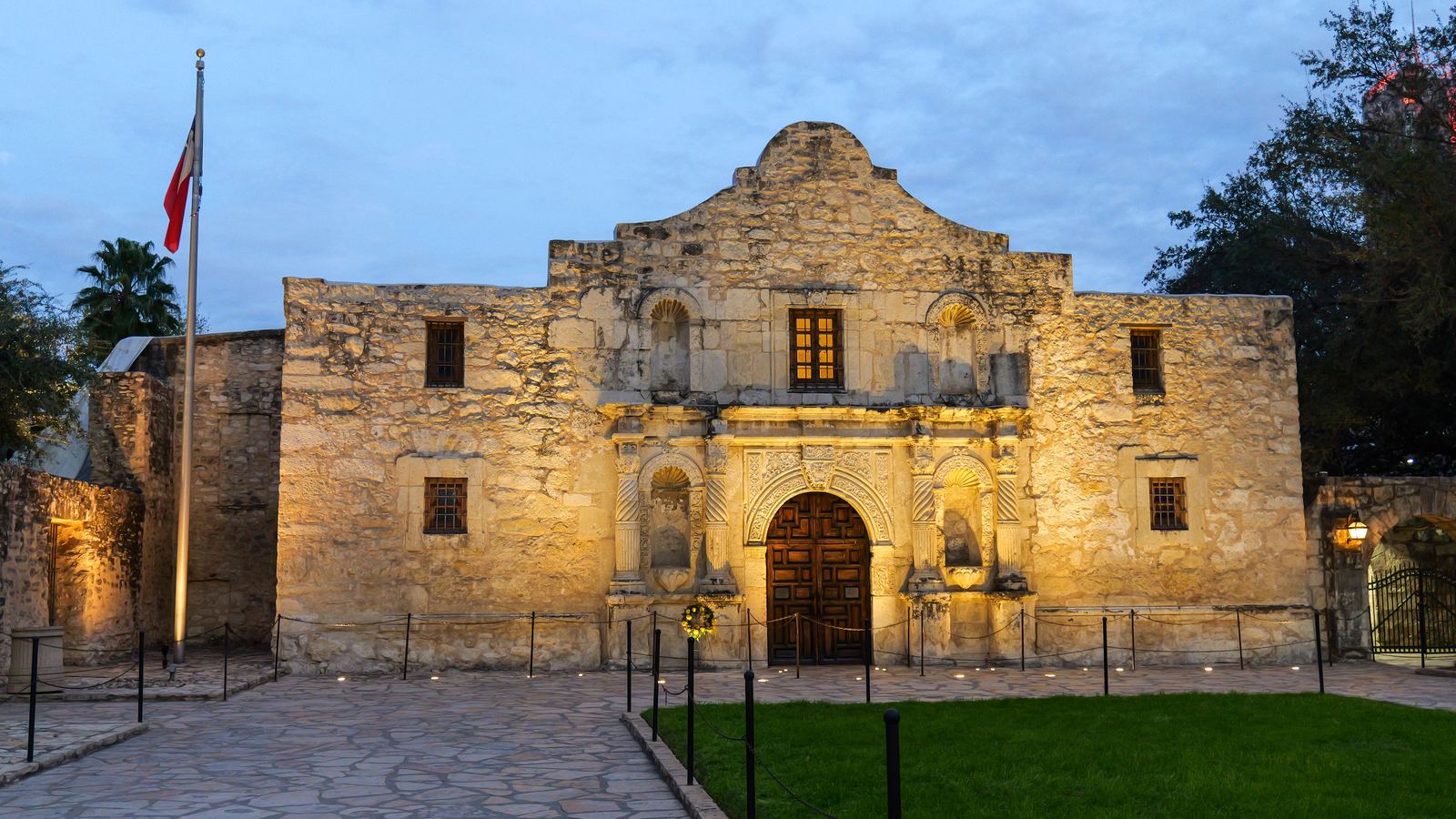 The Alamo floodlit at dusk with the Texas flag flying beside it